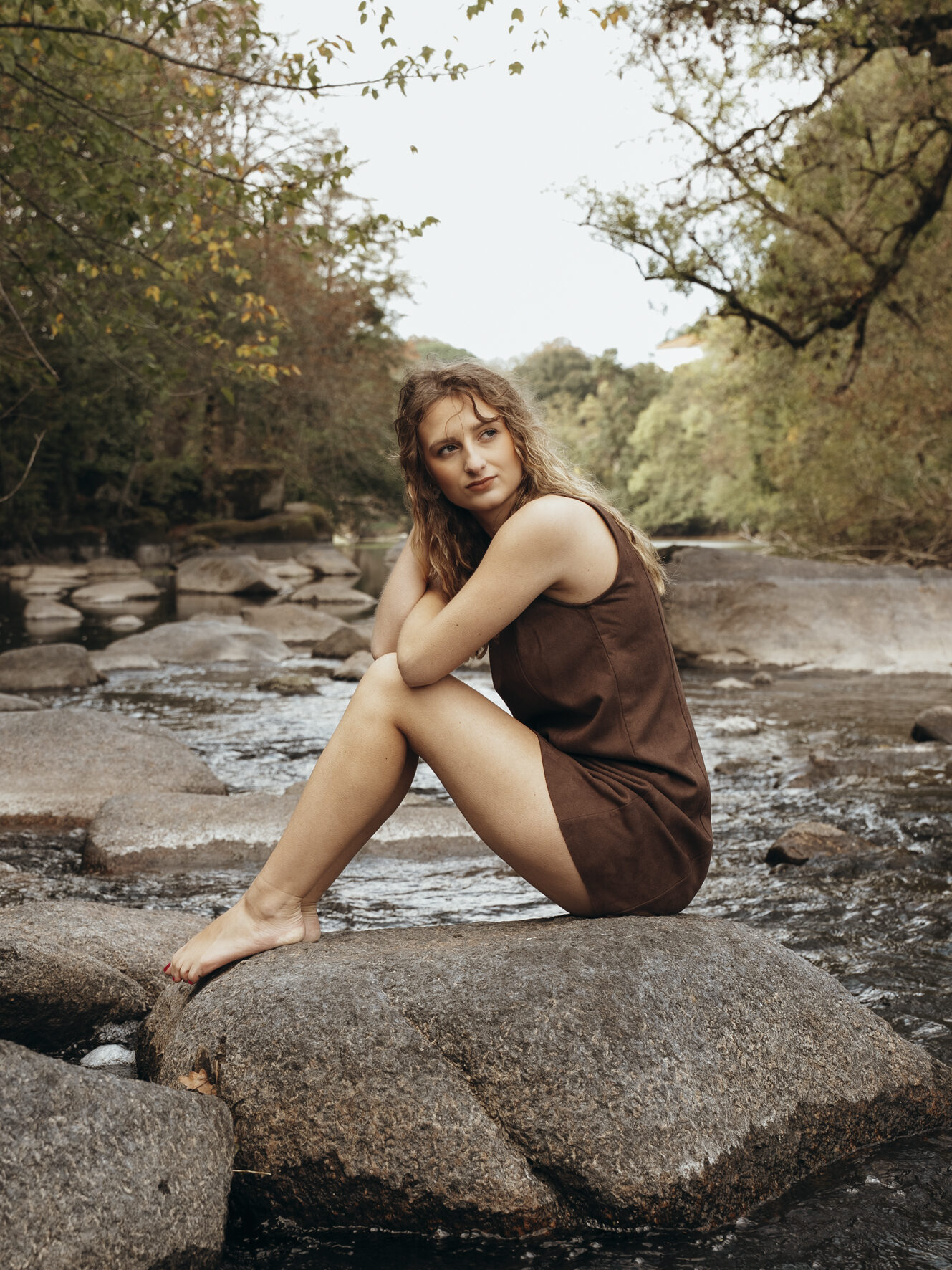 Portrait d’une jeune femme en robe marron, assise sur un rocher au bord d’une rivière
