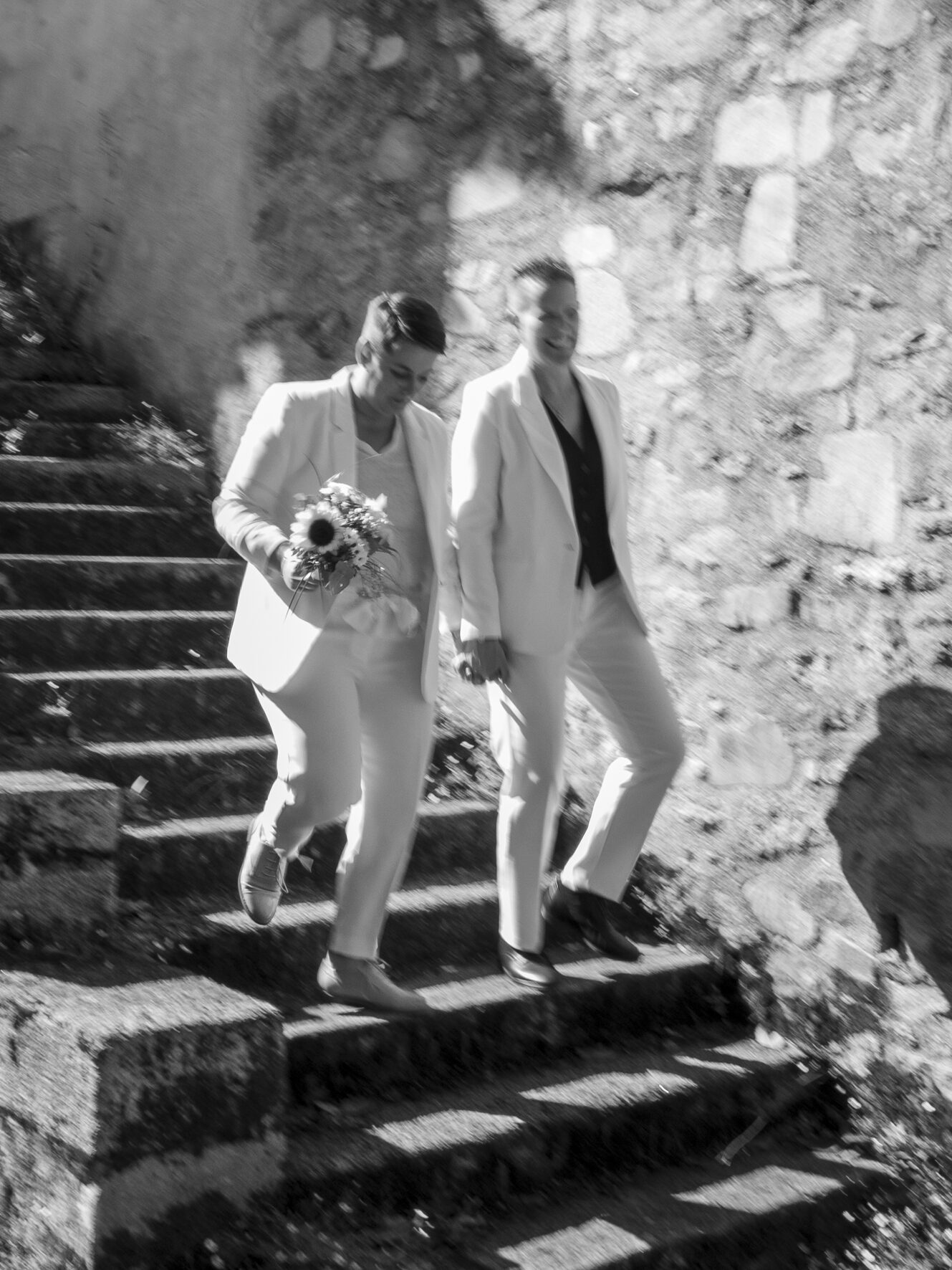 Deux mariées en tailleur blanc descendant un escalier de pierre, bouquet de tournesols à la main, noir et blanc