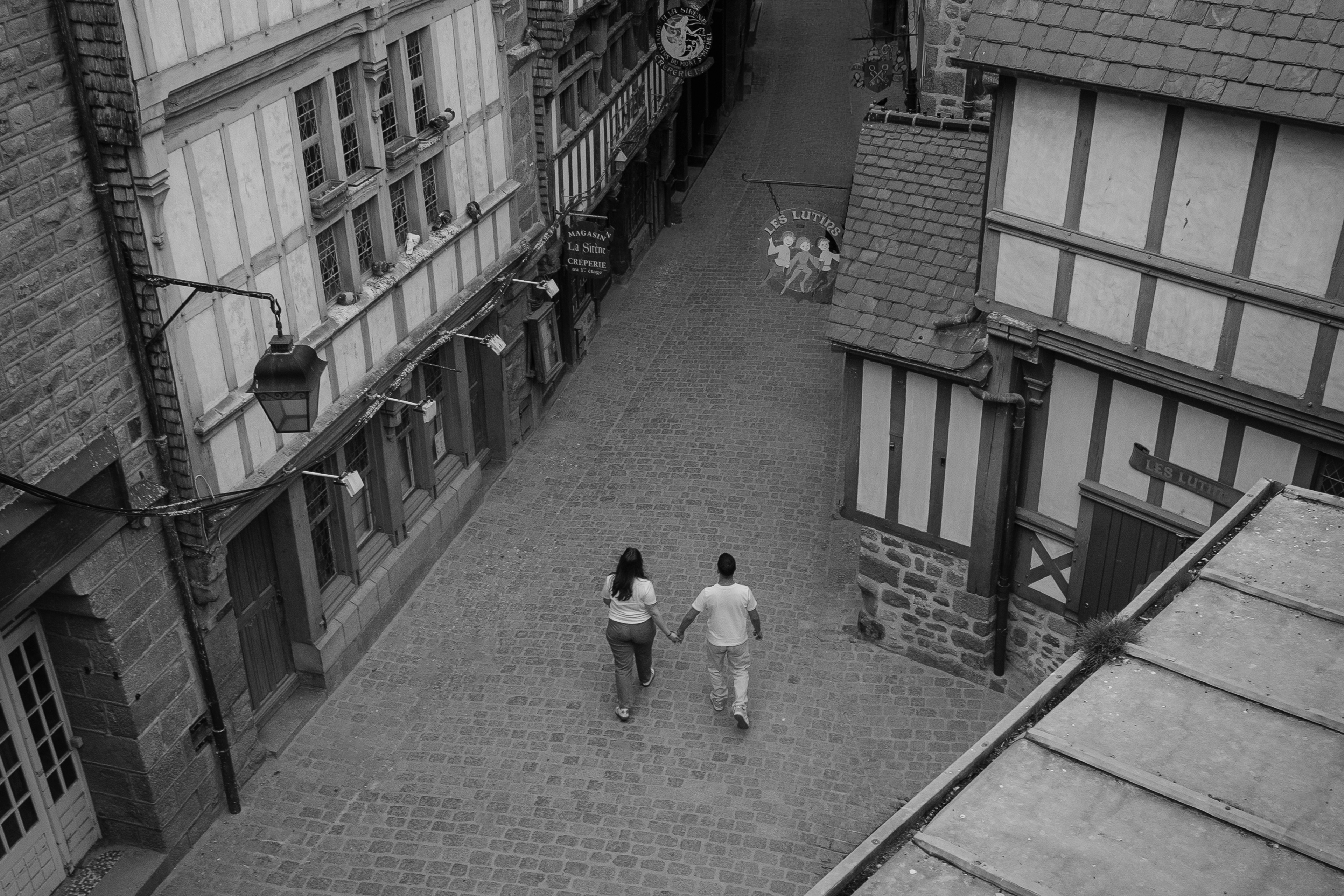 Couple main dans la main dans une ruelle pavée du Mont-Saint-Michel, noir et blanc